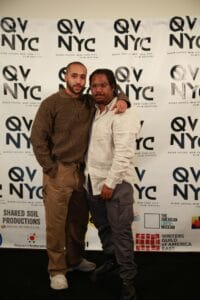 image_168174337 | GLAAD Ronald Hinton (left), wearing all brown, with short brown hair, brown skin and Brandon C. Smith (right), in a white long sleeve with dark pants with a QVNY step and repeat which is in black and white.