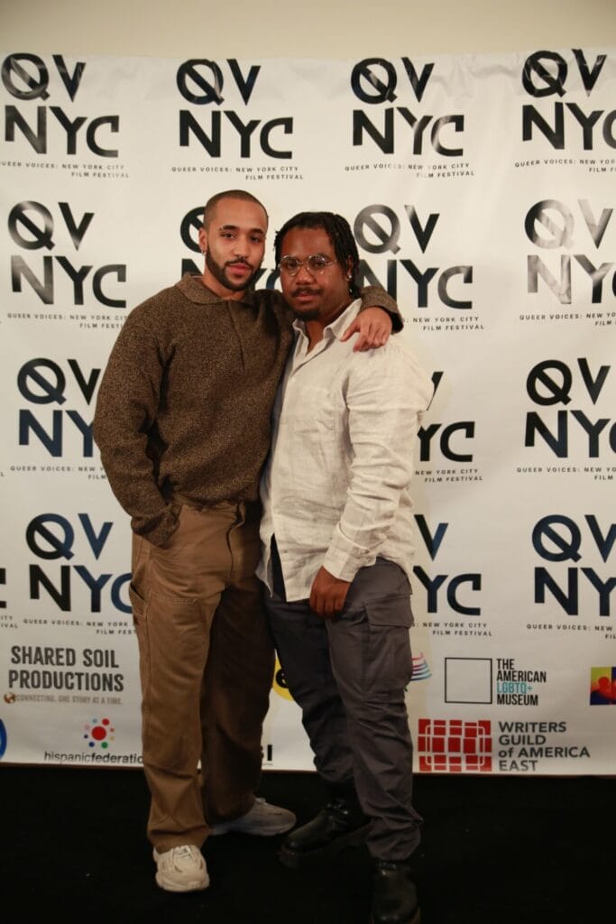 image_168174337 | GLAAD Ronald Hinton (left), wearing all brown, with short brown hair, brown skin and Brandon C. Smith (right), in a white long sleeve with dark pants with a QVNY step and repeat which is in black and white.