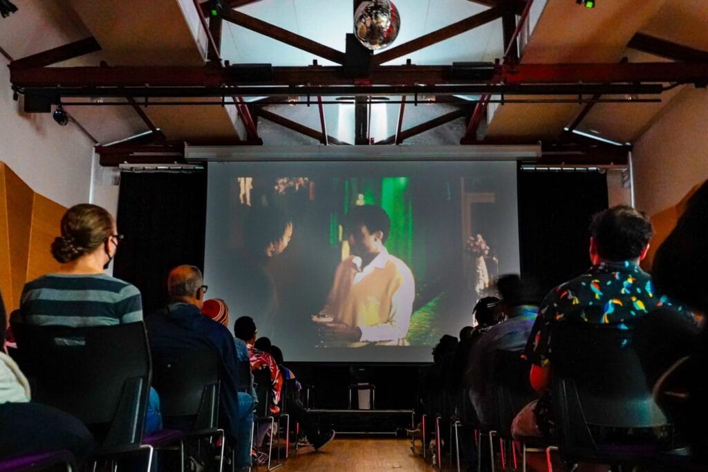 Dark room at The Center on the 3rd floor. A film is playing on a big screen and there are people stacked in seats, with a perfect walk way down toward the screen projecting two people to the left in a dark room with a green light in the background.