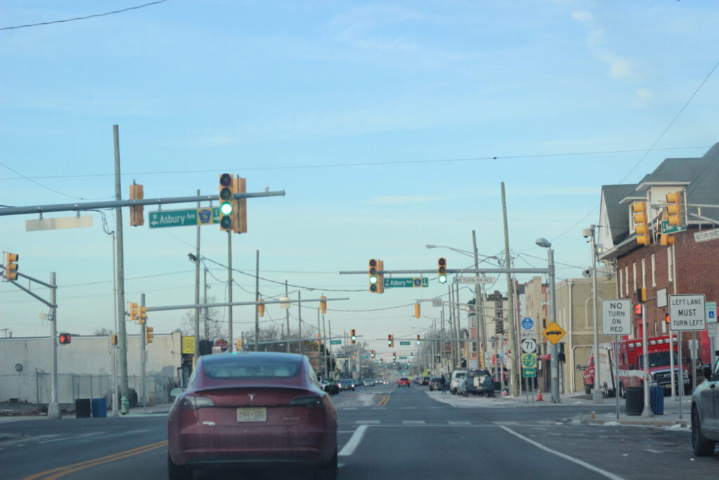 Main Street in Asbury Park NJ. Asbury Ave is ahead of the lights, going green all the way down. The sky is blue, the firestation to the left, and a maroon Tesla is in front of the light. The skyis blue, just before sunset. | GLAAD Main Street in Asbury Park NJ. Asbury Ave is ahead of the lights, going green all the way down. The sky is blue, the firestation to the left, and a maroon Tesla is in front of the light. The skyis blue, just before sunset.