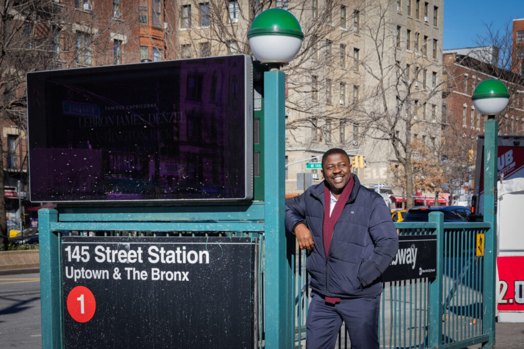 Dannie Dinh 2024x1 | GLAAD Edafe Okporo, candidate for NYC City Council Dis. 7 stands at the 145th St subway station smiling. The subway station is green, and has a title card that reachs "145th Street Station: Uptown & The Bronx, 1 train"