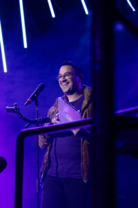 BTB_monday-1 | GLAAD George Strus on stage under blue lights, smiling into the audience with glasses on