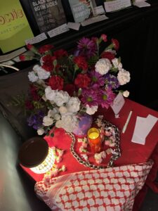 IMG_4737 | GLAAD overhead shot of a small end table with a red table cloth, covered in tributes to Cecilia Gentili. Flowers, a keffiyeh, a chain link necklace, and more