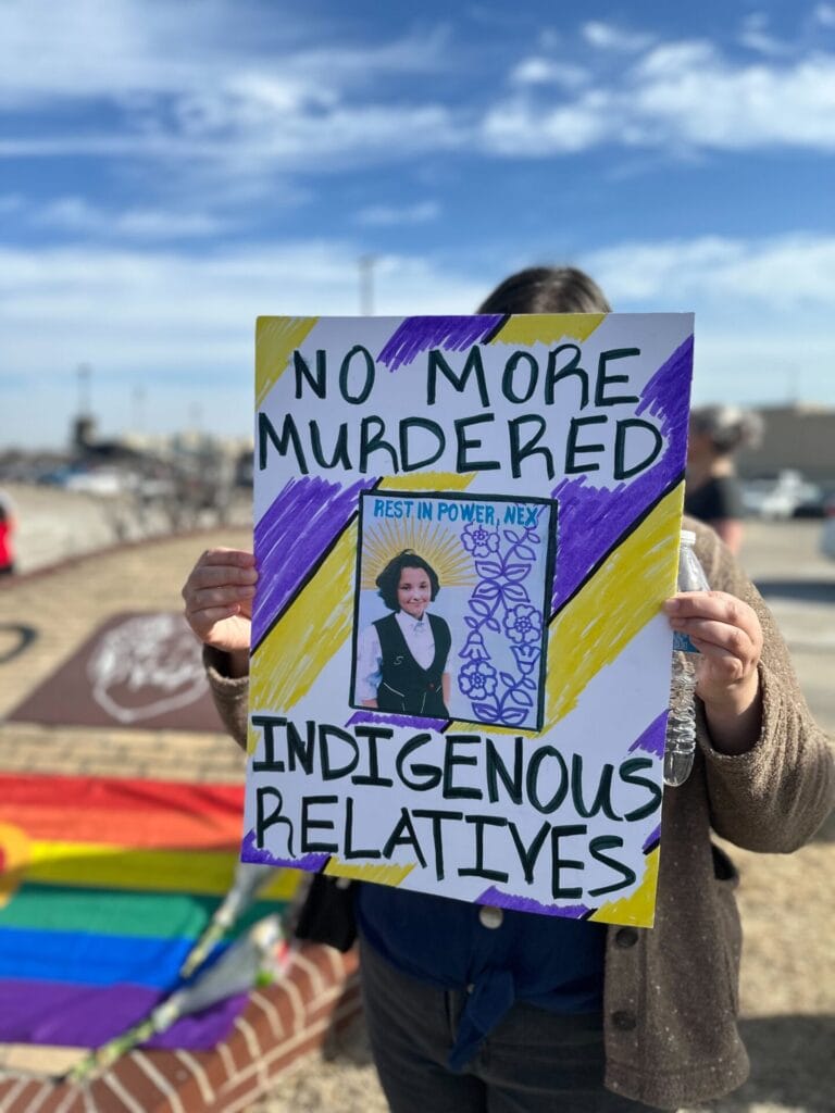 IMG_1837 1 | GLAAD Owasso High School student holds a sign that reads "No More Murdered Indigenous Relatives" outside the high school during a walk out. The sky is blue with scattered clouds, a pride flag lays in the distance where the edges of the photo blur.