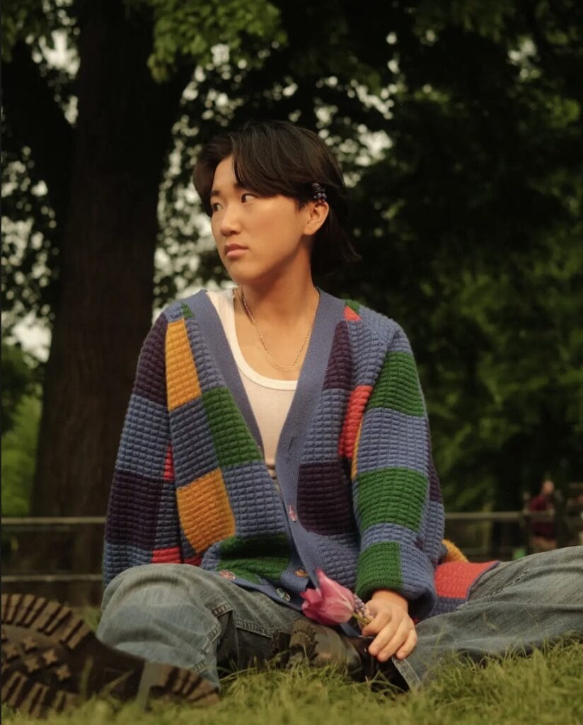 A non-binary trans man sitting on grass, holding a flower | GLAAD A non-binary trans man sitting on grass, holding a flower