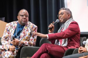 Reginald+3 | GLAAD Reginald Webber, black, in red suit with legs crossed left arm moving as he speaks with a mic close to his face. He speaks on a panel it appears next to a man, black and bald, with black glasses in a rainbow, floral pattern suit and a blue shirt.