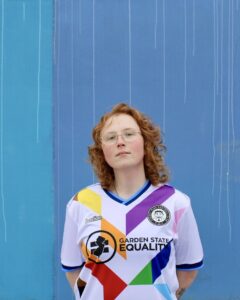 aedy+2 | GLAAD Aedy Miller, white, red, wavy hair wears glasses and a white and rainbow designed Garden State Equality jersey infront of a blue wall.