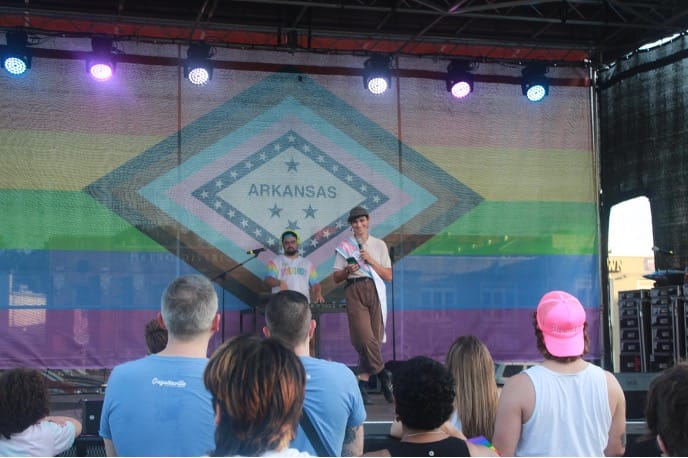 Two people on an outdoor stage speaking into microphones in front of a large rainbow Pride banner that reads Arkansas, with an audience watching.",