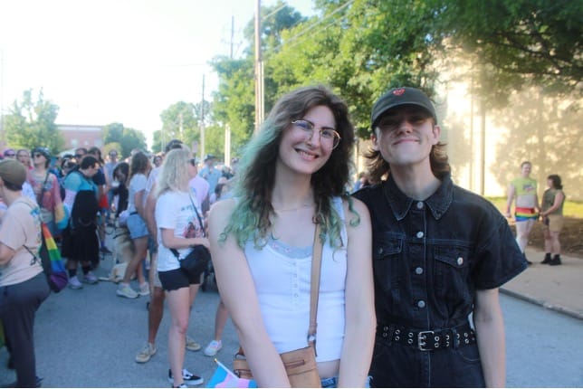 Two young friends pose and smile at a sunny street parade with a crowd in the background, one with green-tinted hair and the other in a black denim outfit.