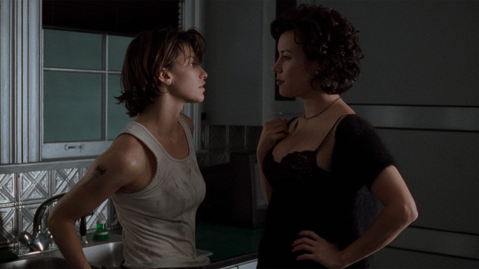 Bound Shot | GLAAD Two women stand close in a dim kitchen, one in a dirty white tank top and the other in a black lace dress, facing each other in a tense stare down.