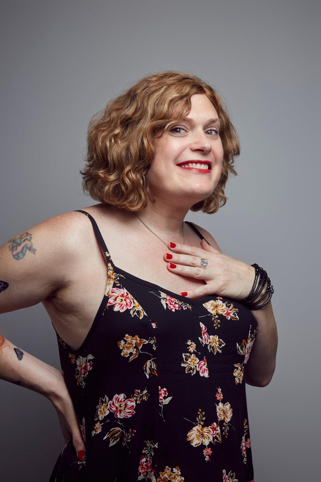 lilly wachowski headshot | GLAAD Smiling woman with short curly blonde hair in a floral dress, hand on chest, gray studio background