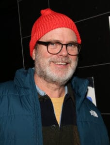 Pria tersenyum dengan beanie rajut merah cerah, kacamata berbingkai gelap, dan janggut abu-abu dengan jaket biru.