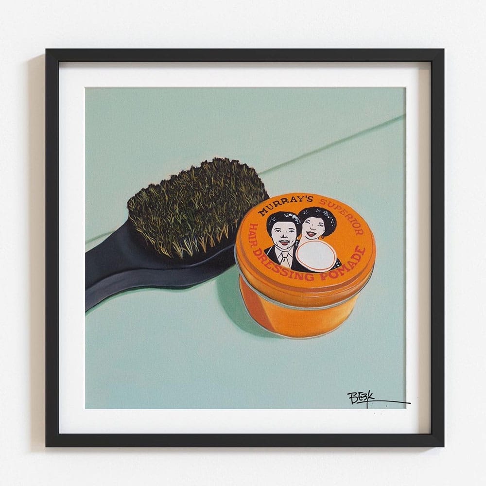 Framed still-life painting showing a black-handled hairbrush beside an orange jar labeled 'Murray's Superior Hair Dressing Pomade' on a pale blue-green backdrop.
