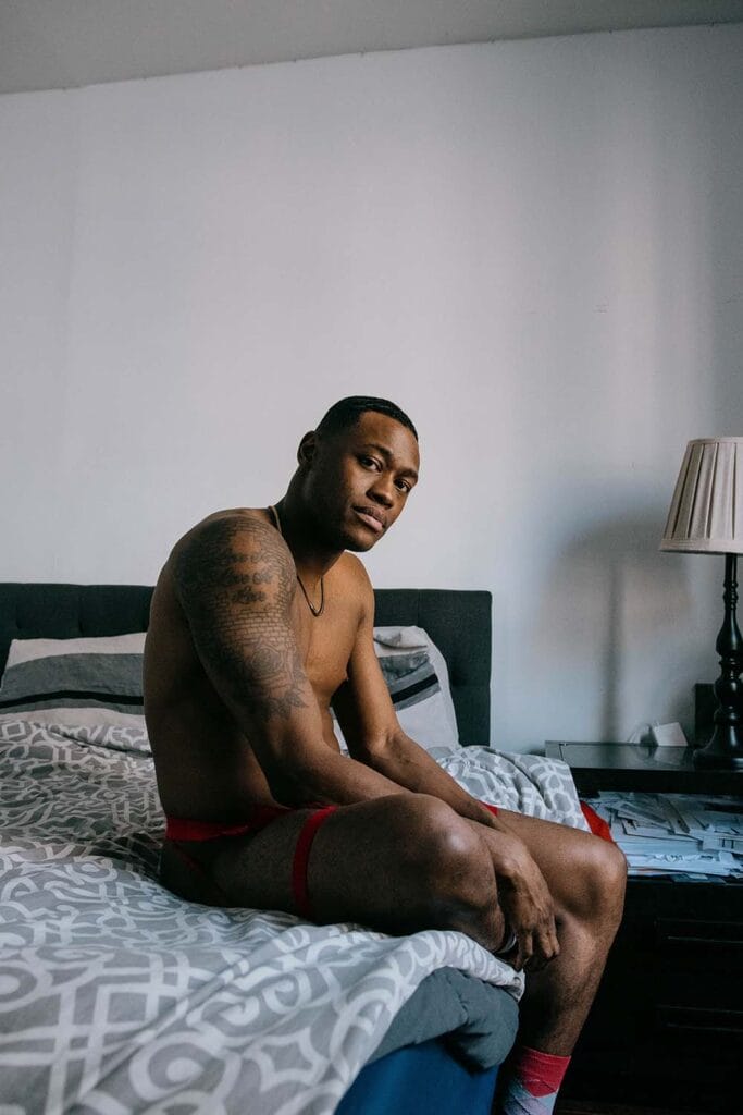 Shirtless man with a large arm tattoo sits on a bed, facing the camera in a bedroom with a bedside lamp.