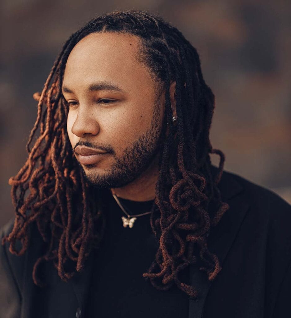 Portrait of a man with long brown dreadlocks wearing a black shirt, looking to the left.