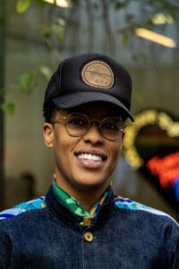 Smiling person wearing a black cap with a tan circular logo, round glasses, and a denim jacket with a colorful scarf outdoors.