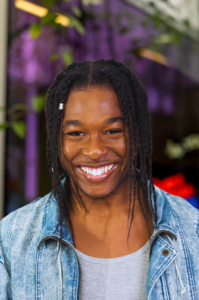 Photograph of the artist Travion Payne | GLAAD Smiling man with dreadlocks wearing a light denim jacket and gray shirt, outdoors.