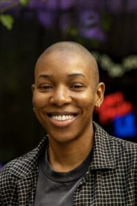 Close-up of a smiling person with a shaved head, wearing a black-and-white checkered jacket and hoop earring.
