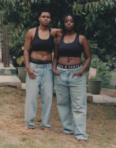 Two women stand side by side outdoors, wearing black sports bras and light jeans, barefoot on grass under a tree, one resting her hand on the other's shoulder.