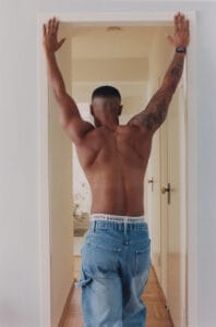 Back view of a shirtless man with arms raised, wearing blue jeans and a white FENTY SAVAGE waistband, standing in a doorway with tattoos on his right arm.