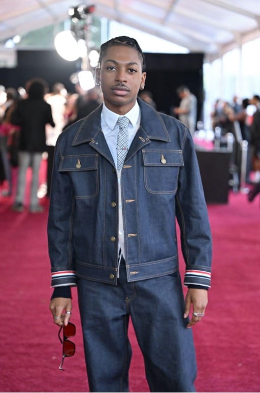 Young man on a pink carpet wearing a denim jacket and jeans with a light blue shirt and patterned tie, holding sunglasses, at a fashion event.