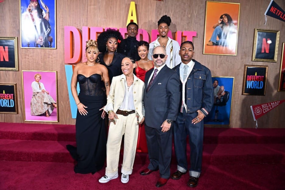 Group of eight people posing on a red carpet at a premiere, wood-paneled backdrop with framed posters and bold pink lettering behind them.