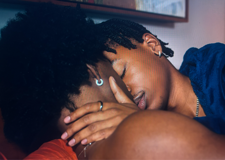 Two people embrace for a kiss, one with short twists and hoop earrings resting on the other's shoulder.