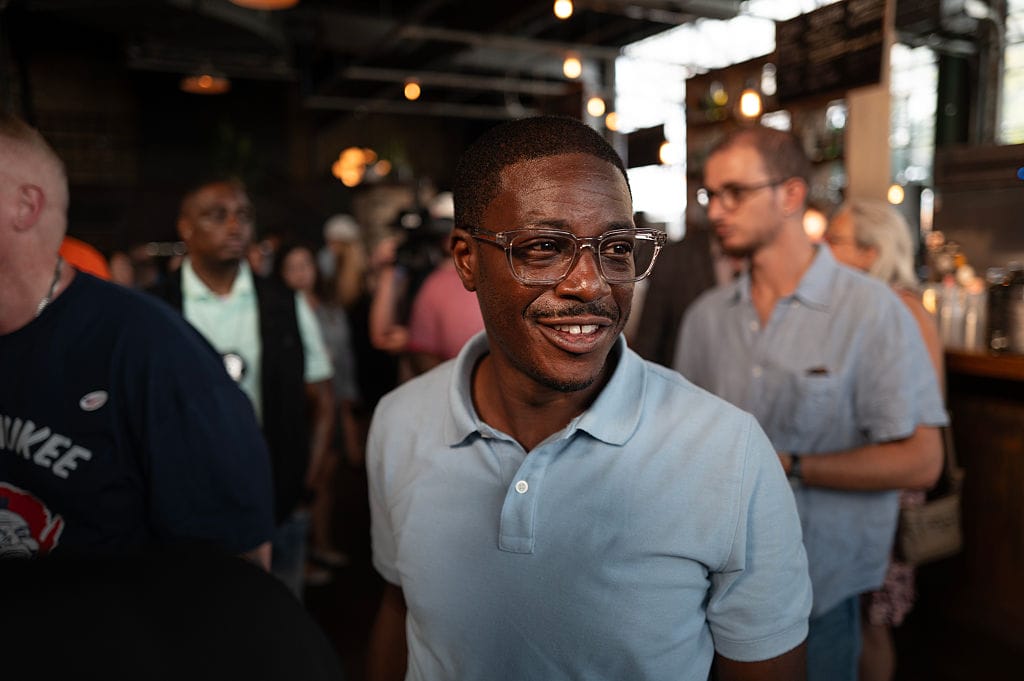 Wisconsin Gubernatorial Candidate David Crowley Holds Campaign Rally On Eve Of Primary Election | GLAAD Smiling man wearing clear glasses and a light blue polo at a crowded indoor social event or bar setting.