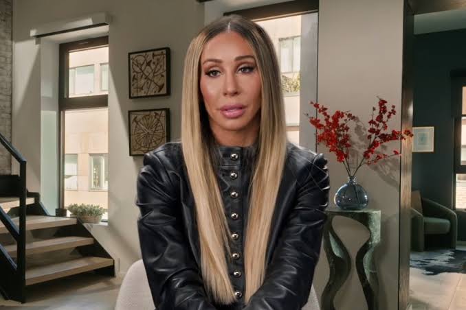 Woman with long straight blonde hair wearing a black leather jacket sits in a modern interior, looking at the camera.