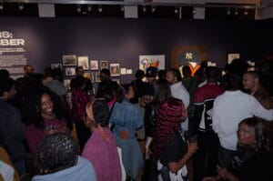 Crowded gallery scene with people viewing framed photos on a blue wall and colorful artwork, conversations ongoing beneath track lighting.