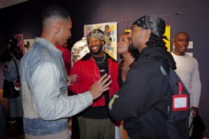 Group of people chatting at an art event, with a man in a blue denim jacket on the left and others nearby against a purple wall with colorful artwork in the background.