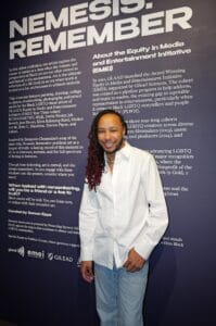 Man with long dreadlocks and a white shirt posing in front of a dark purple wall with large white text art installation. read as backdrop about Nemesis: Remember.