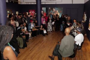 Diverse audience gathered in a gallery, seated and standing, watching a presentation with colorful paintings on dark walls behind them.