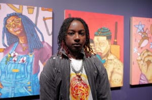 Portrait of a person with braided hair and a nose ring standing in front of colorful gallery paintings far left to right.