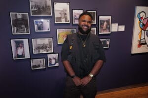 Smiling man with a beard stands in front of a purple gallery wall filled with framed black-and-white photos and a colorful mural on the right.