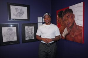 Smiling man in a white T-shirt and cap stands in an art gallery, arms folded, with framed sketches on a blue wall behind him and a large red-framed portrait to his right.