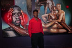 Young man in a red shirts stands before a large surreal mural of intertwined nude figures and faces in bold colors.
