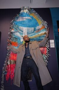 Man posing dramatically in front of a large colorful fiber-art sculpture, wearing a beige trench coat and dark jeans.