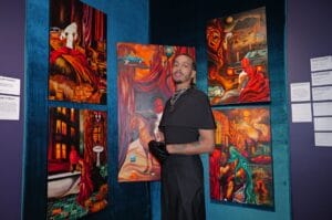 Man in black clothing with gloves posing in an art gallery, standing before bold, surreal paintings on teal walls.