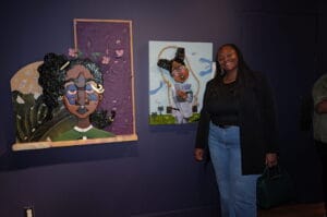 Woman in a black top and jeans stands beside two colorful art pieces on a purple wall, smiling at the camera.