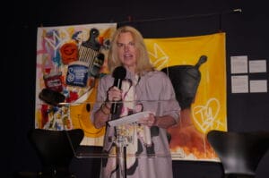 Woman in a lavender blouse speaks into a handheld microphone at a clear lectern on a stage, with a colorful mural backdrop behind her.