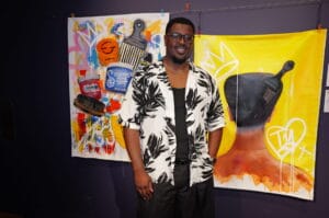 Man in a black-and-white tropical shirt posing in front of large, colorful art banners with a paintbrush motif and bold yellow background.