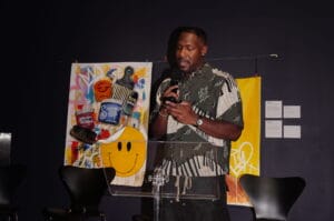 Man speaking into a handheld microphone at a clear podium in front of a colorful art backdrop.