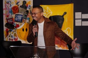 Man in brown jacket speaks into a microphone at a clear podium, gesturing with his right hand in front of colorful graffiti art banners.