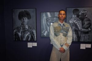 Young man poses in front of grayscale armor portraits in a gallery, wearing a light denim jacket and beige pants. A wall with framed artworks and small description plaques is visible behind him.