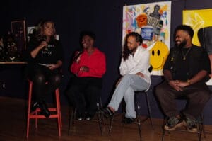 Panel discussion: four people sit on stools on a stage, microphones in hand, colorful art posters in the background.