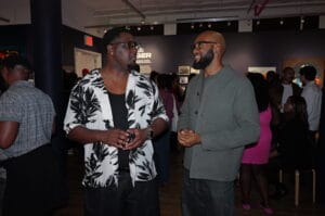 Two men in discussion at an art gallery, with other guests mingling in the background