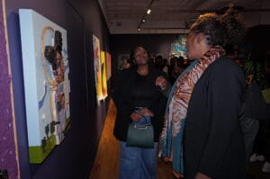 Two women in a gallery examine colorful wall art; one in a black blazer smiles, holding a green handbag, while the other wears a patterned scarf.