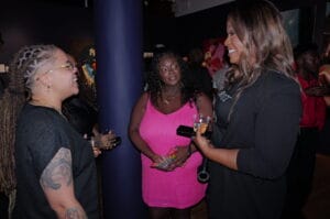 Three women in a social setting chat and laugh, holding drinks; one wears a pink dress, another with tattoos and glasses, a third in black with long hair.