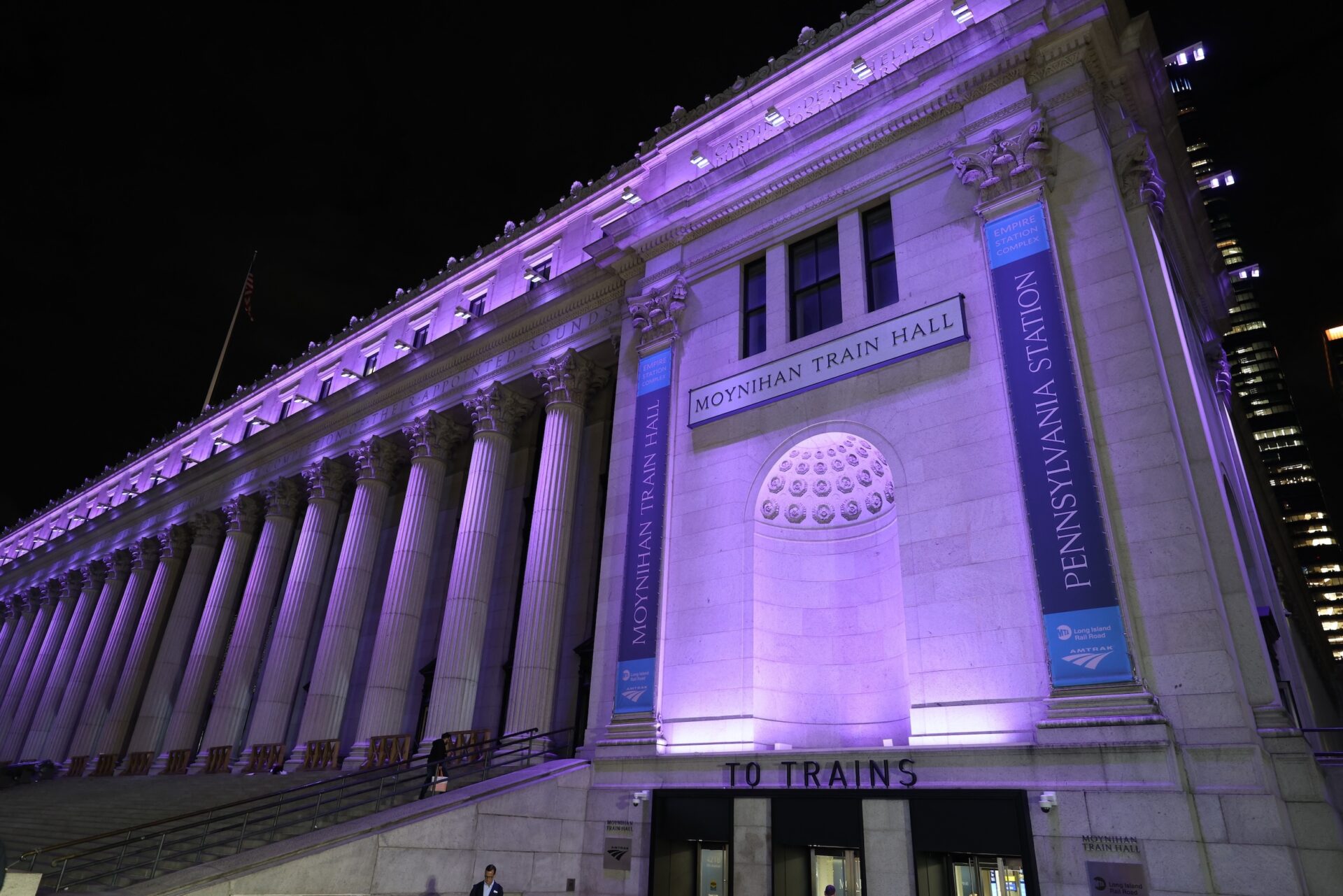 Moynihan Train Hall exterior at night with purple lighting, tall Corinthian columns, and banners along the façade.